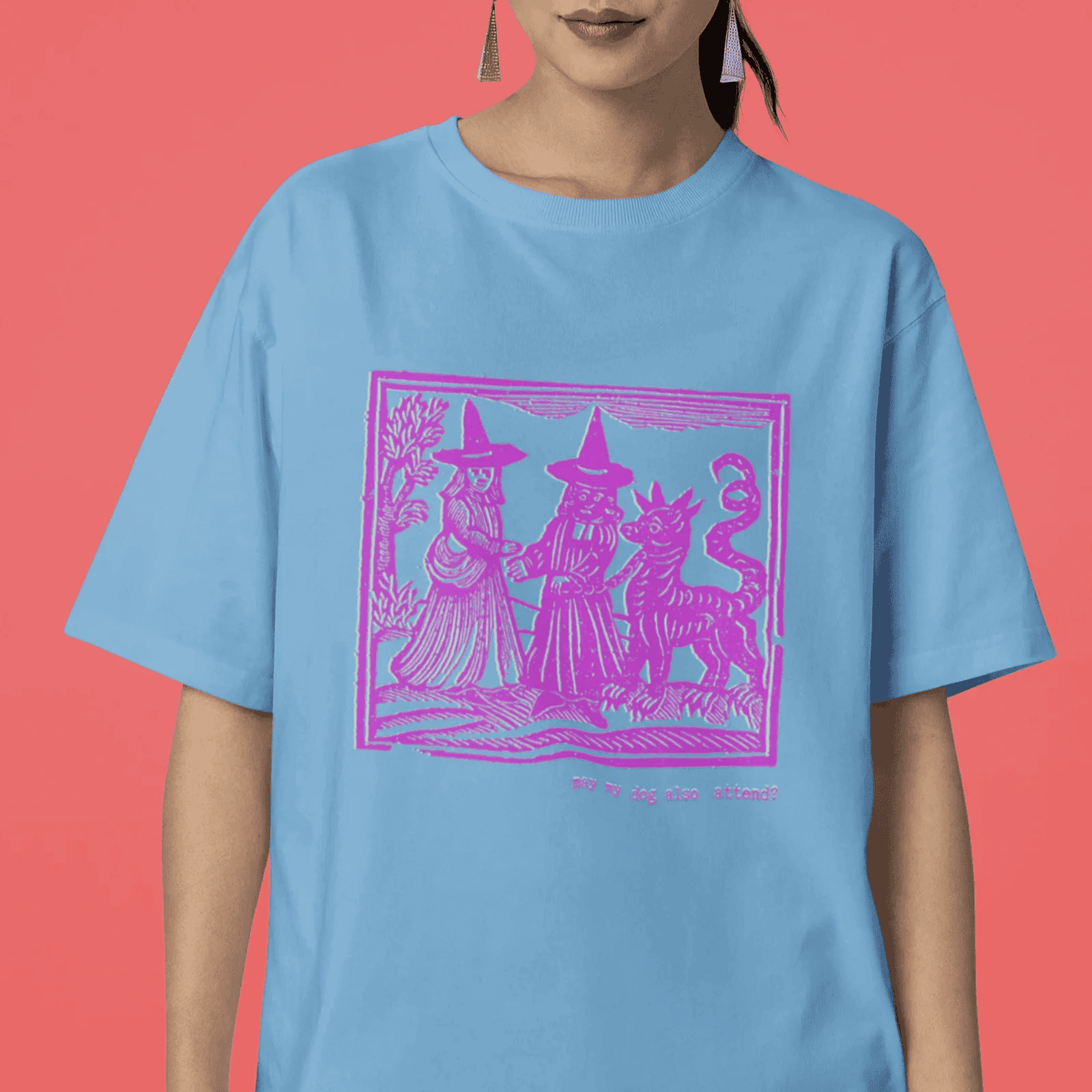 Woman wearing carolina blue staple t-shirt with hot pink medieval woodcut style design of witches with a demon-dog saying "ma