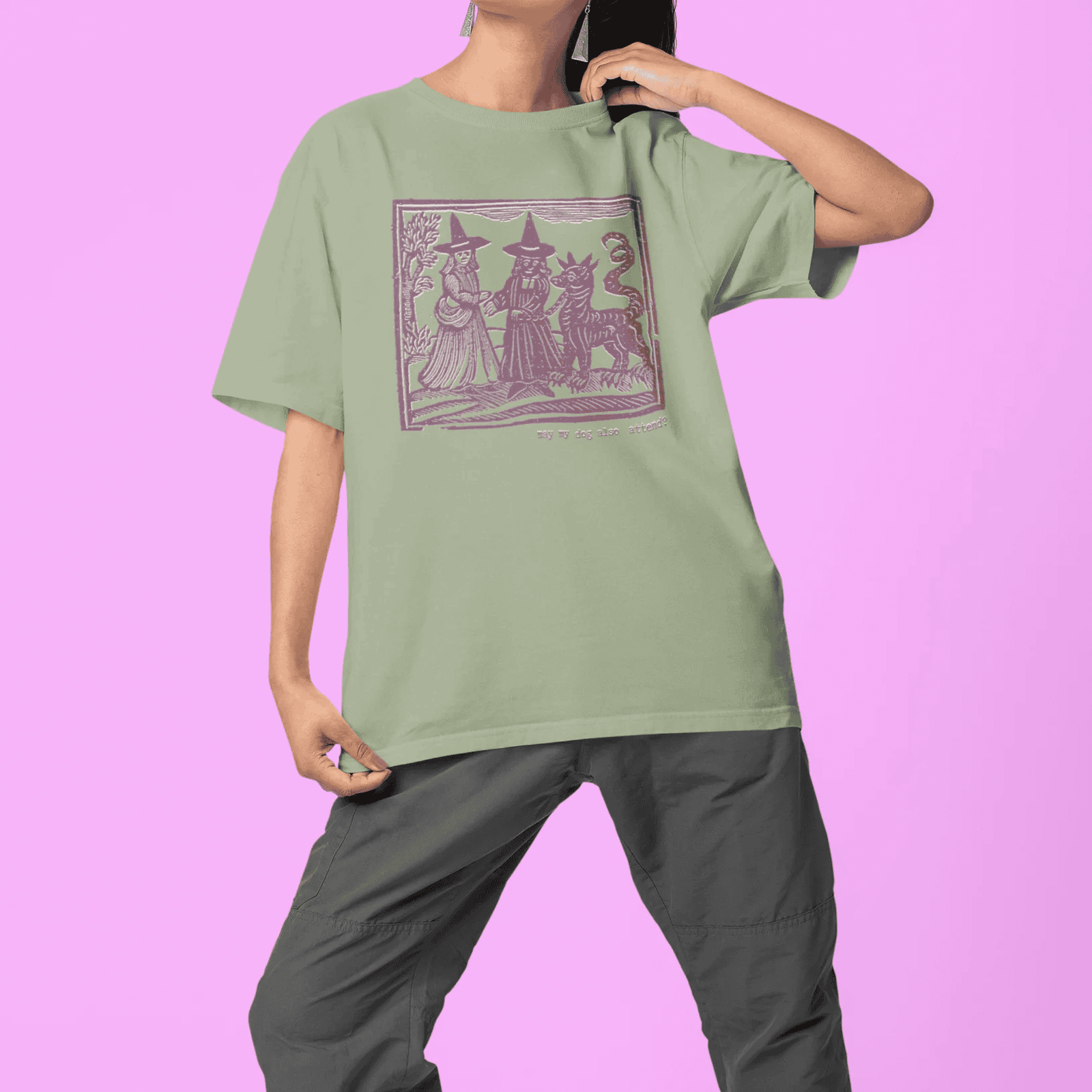 Woman wearing sage classic t-shirt with maroon design in medieval woodcut style design of witches with a demon-dog saying "ma