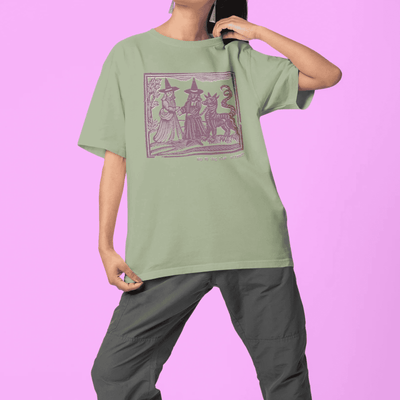 Woman wearing sage classic t-shirt with maroon design in medieval woodcut style design of witches with a demon-dog saying "ma